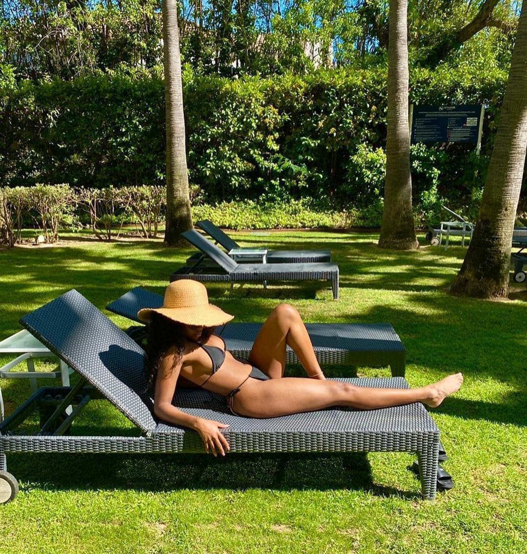 A woman in a straw hat and dark bikini reclines on a woven lounge chair on a grassy lawn near palm trees.