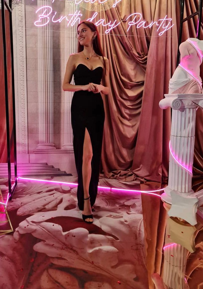 A woman stands in a black strapless gown with a high slit before a pink-lit backdrop featuring a neon sign.