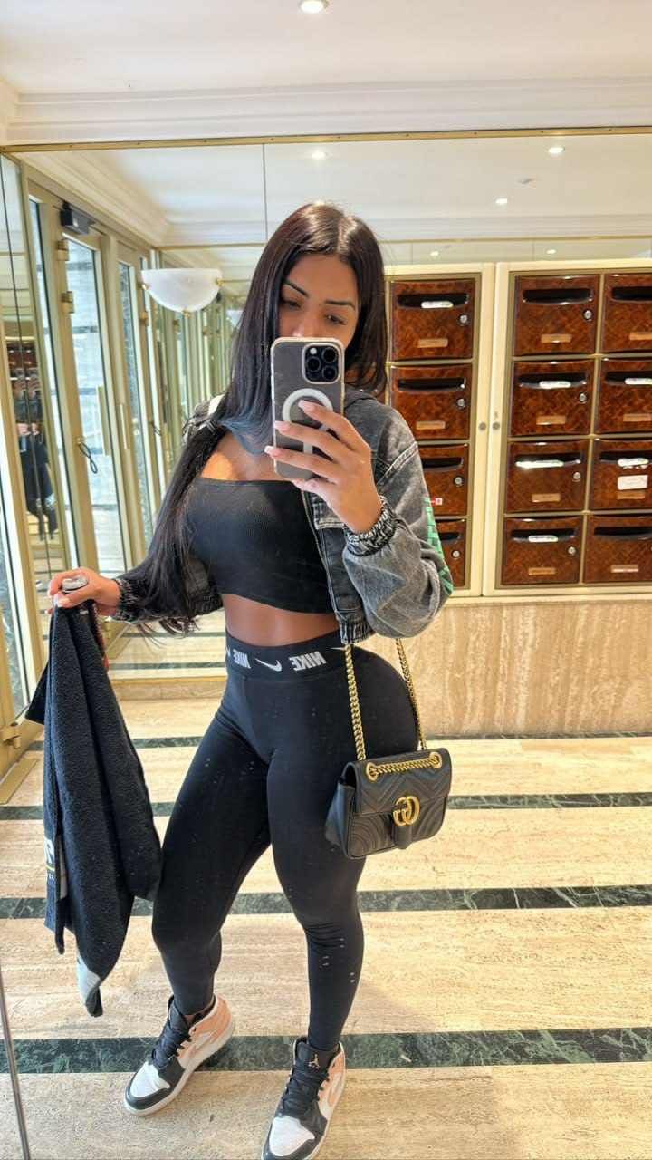 A woman takes a mirror selfie in a hotel lobby wearing a black crop top, leggings, denim jacket, and sneakers, holding a towel.