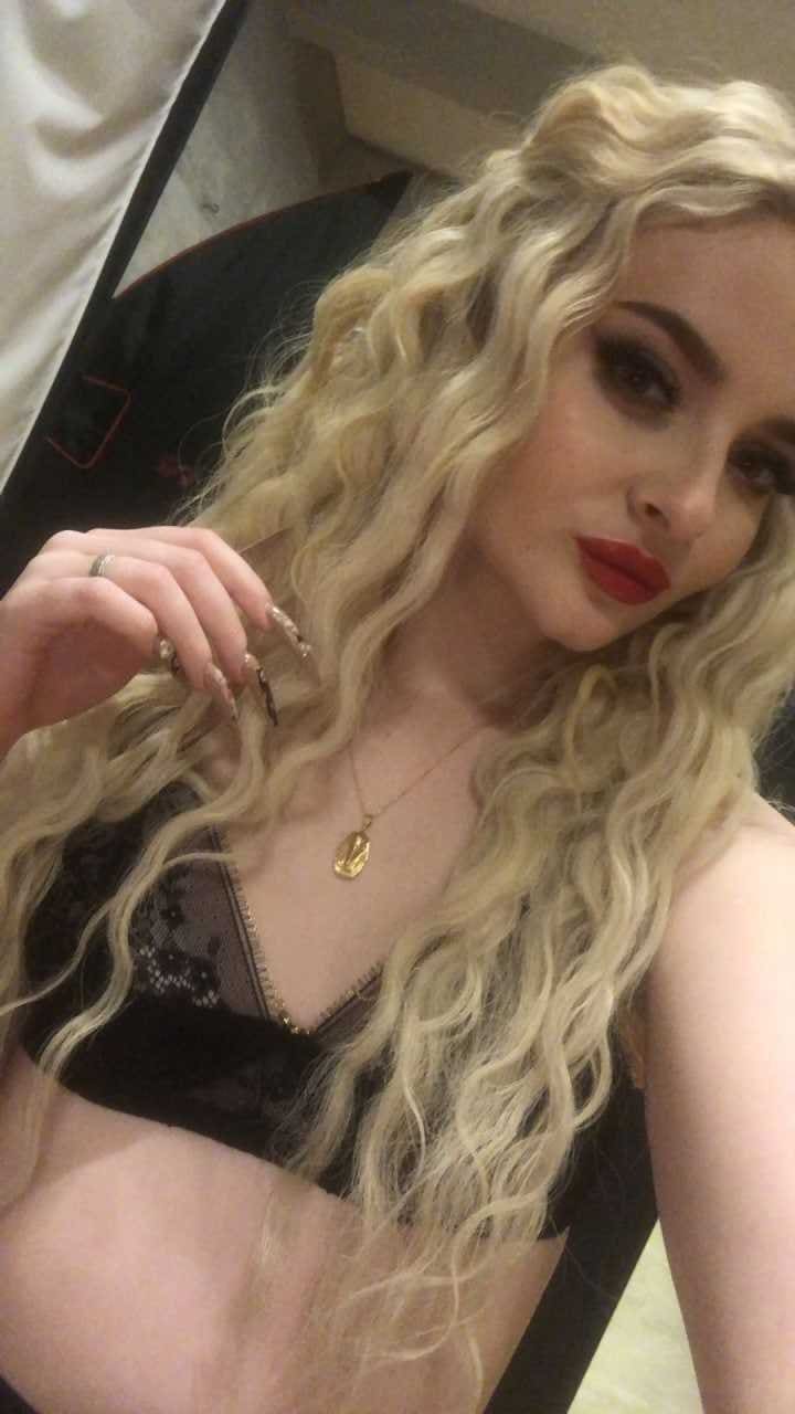 Woman in black lace bra holding a strand of hair near her face, with dark eye makeup in an indoor setting.