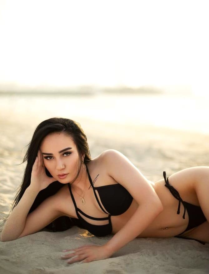 A woman lies on her side on a sandy beach in a black string bikini and thin necklace, resting near her head.