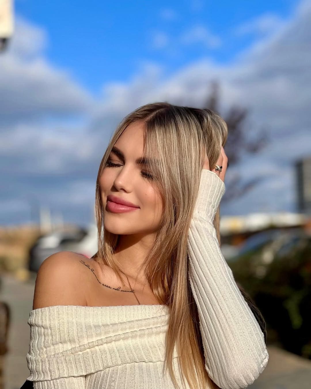 A woman with long blonde hair stands outdoors under a blue sky, wearing a cream ribbed off-the-shoulder sweater with one hand behind her head.