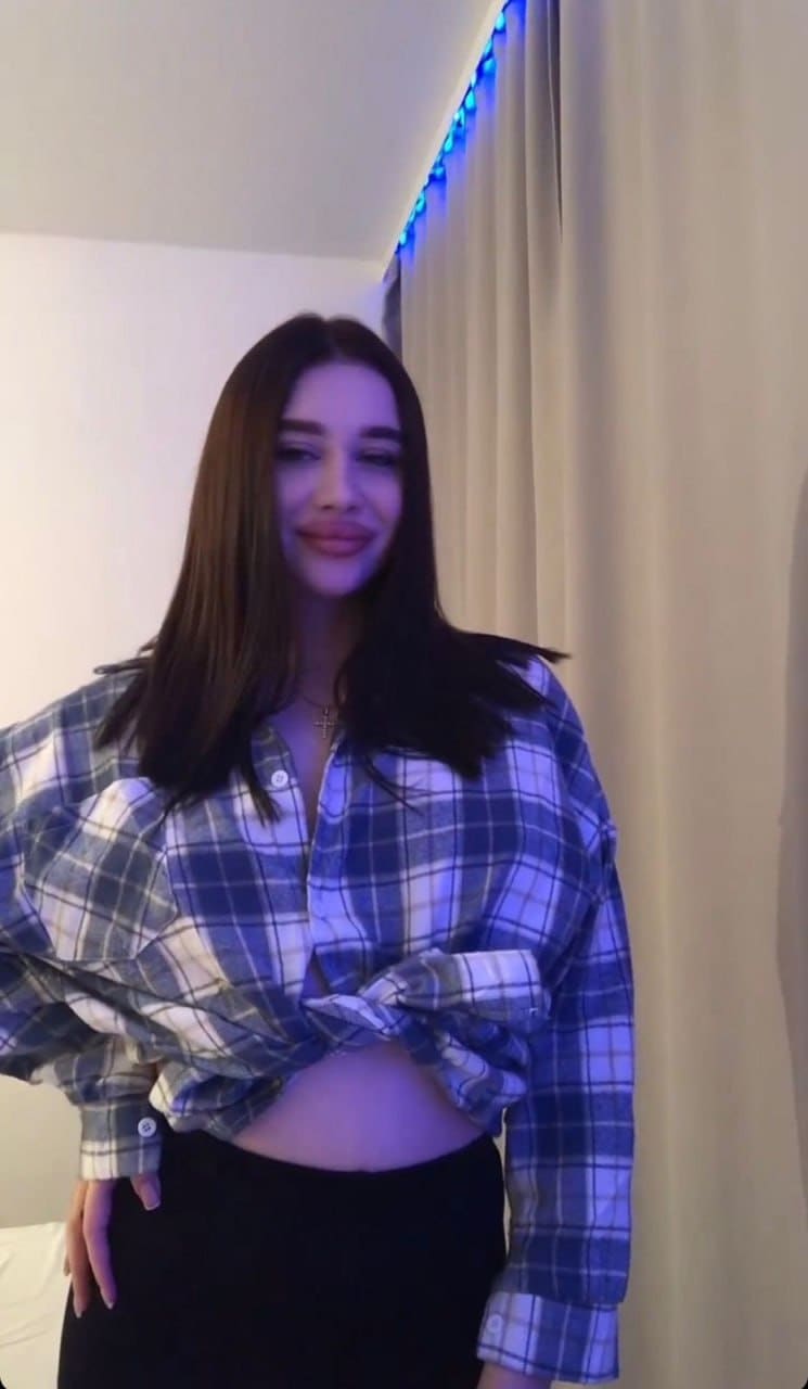 Angelina stands indoors in a blue plaid shirt tied at the waist and black pants, posing against a curtain under blue lighting.
