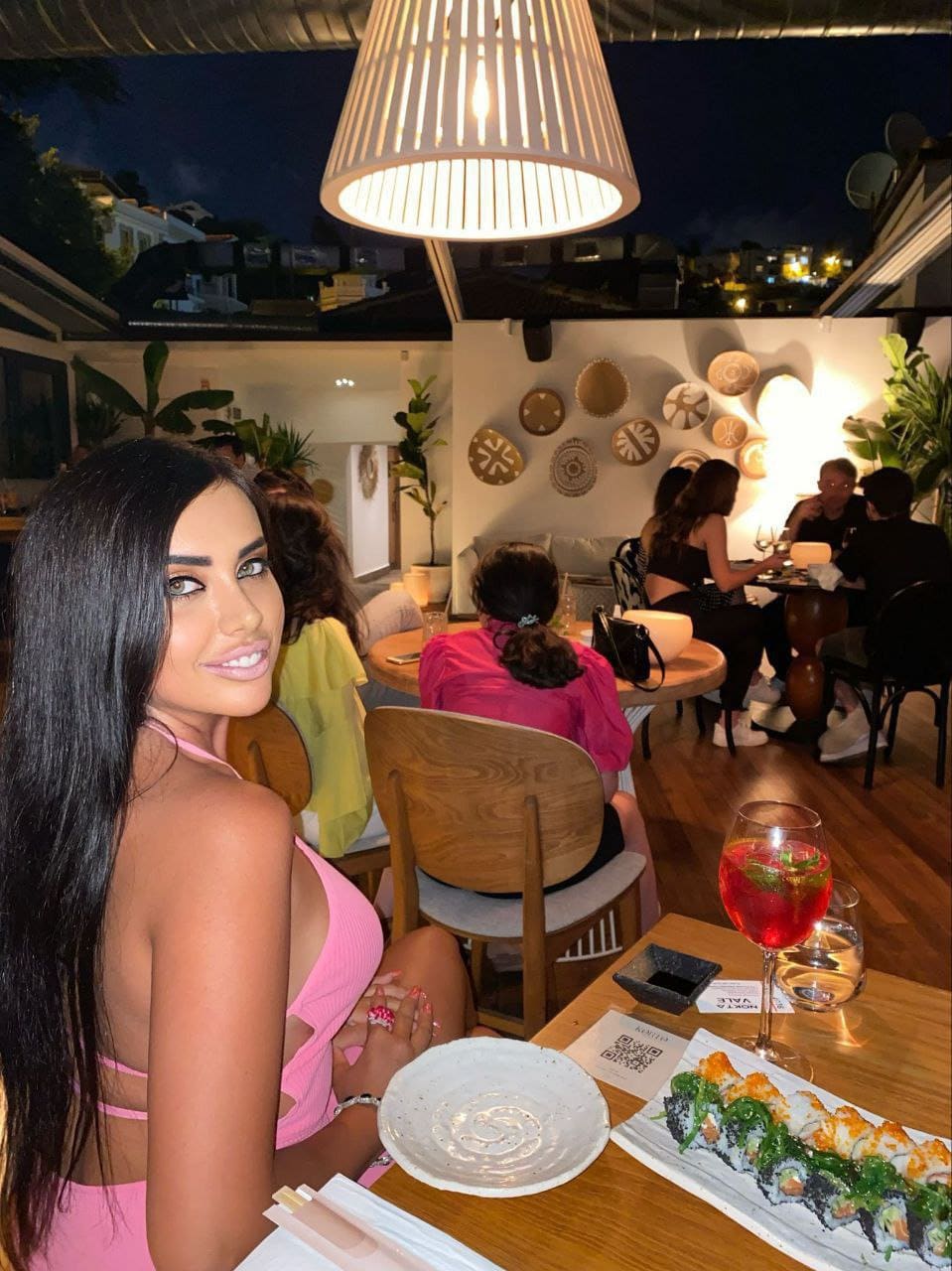 A woman in a pink sleeveless top with a low back sits at a wooden table with sushi and a red drink in a restaurant setting.