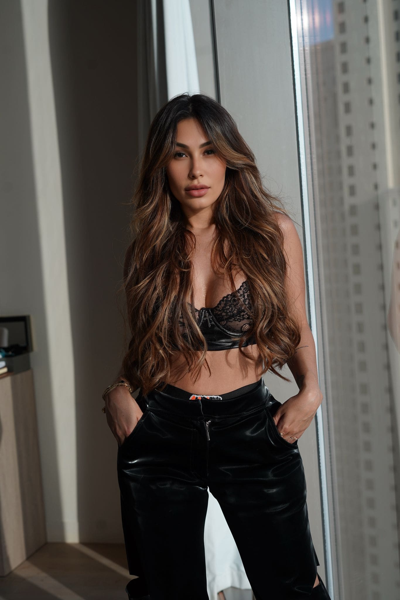 A woman stands indoors near a window in a black lace bralette and patent leather pants, hands in pockets, with bare midriff.