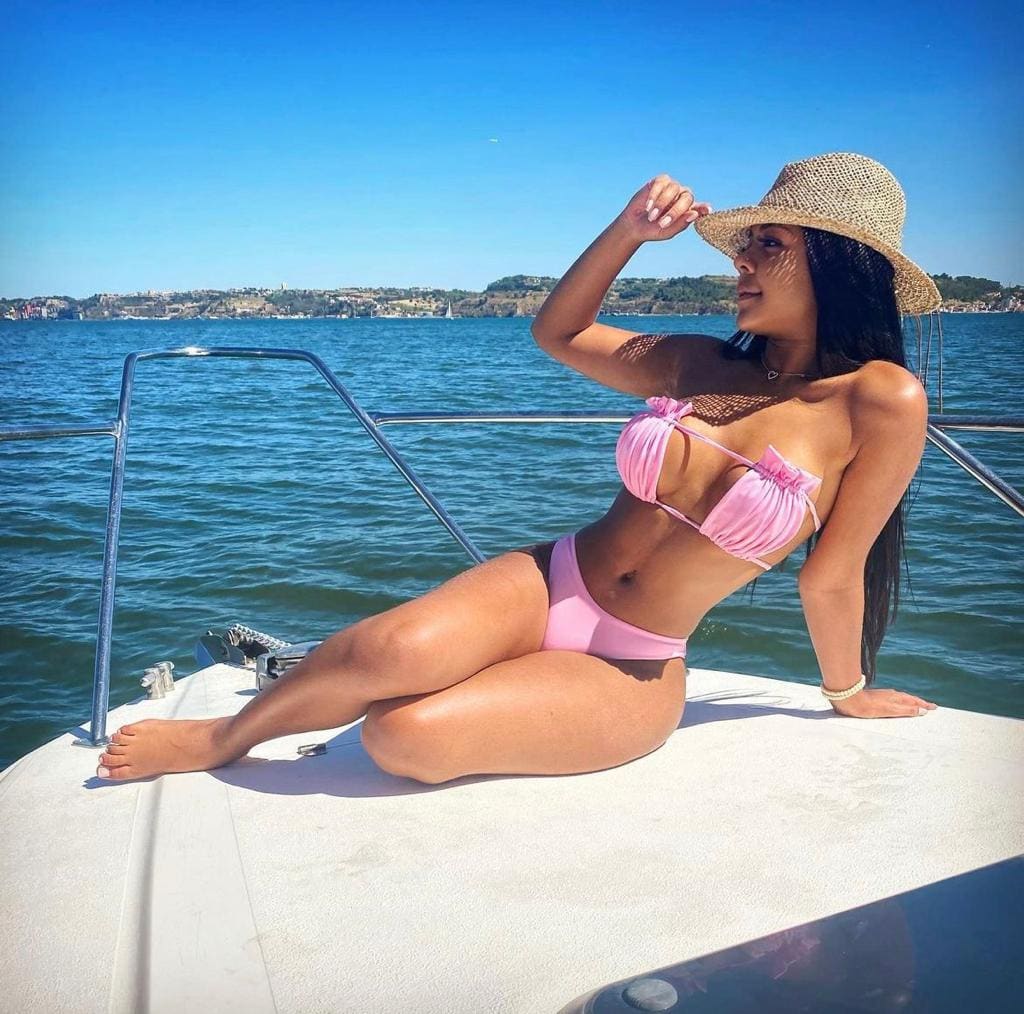 A woman in a pink bikini and straw hat sits on a boat's bow, hand on the brim, against open water and a distant shoreline.