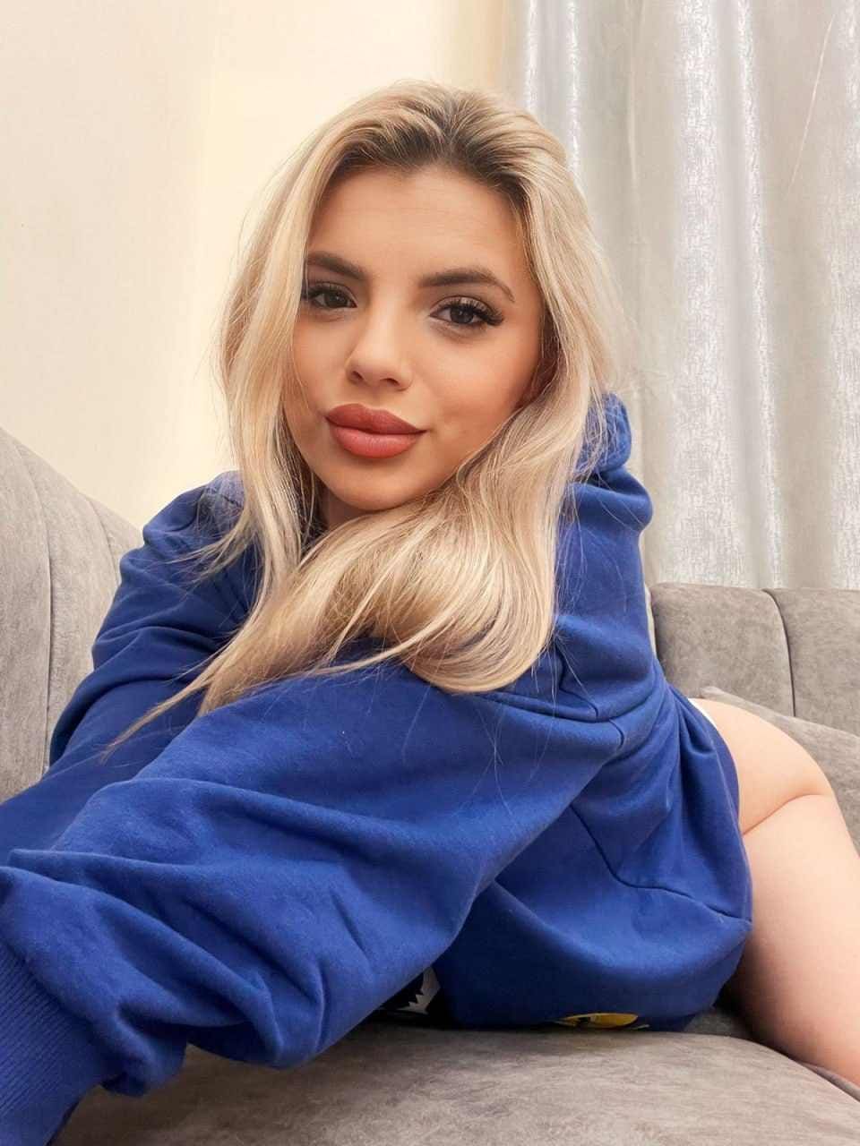Juliette sits on a gray couch in an oversized blue hoodie, bare legs visible, with a light curtain behind her.