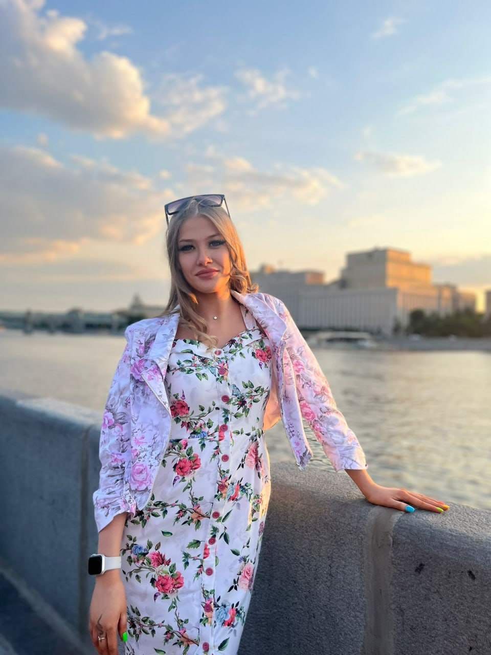 Dana stands by a railing near water in a white floral dress and jacket, with a city skyline behind her.