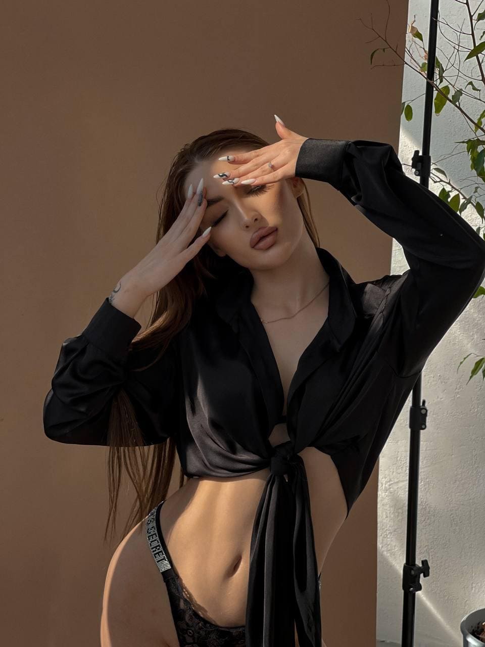 A woman with brown hair stands outdoors against a tan wall in a black satin shirt and lace underwear, hands raised to her forehead near a plant.
