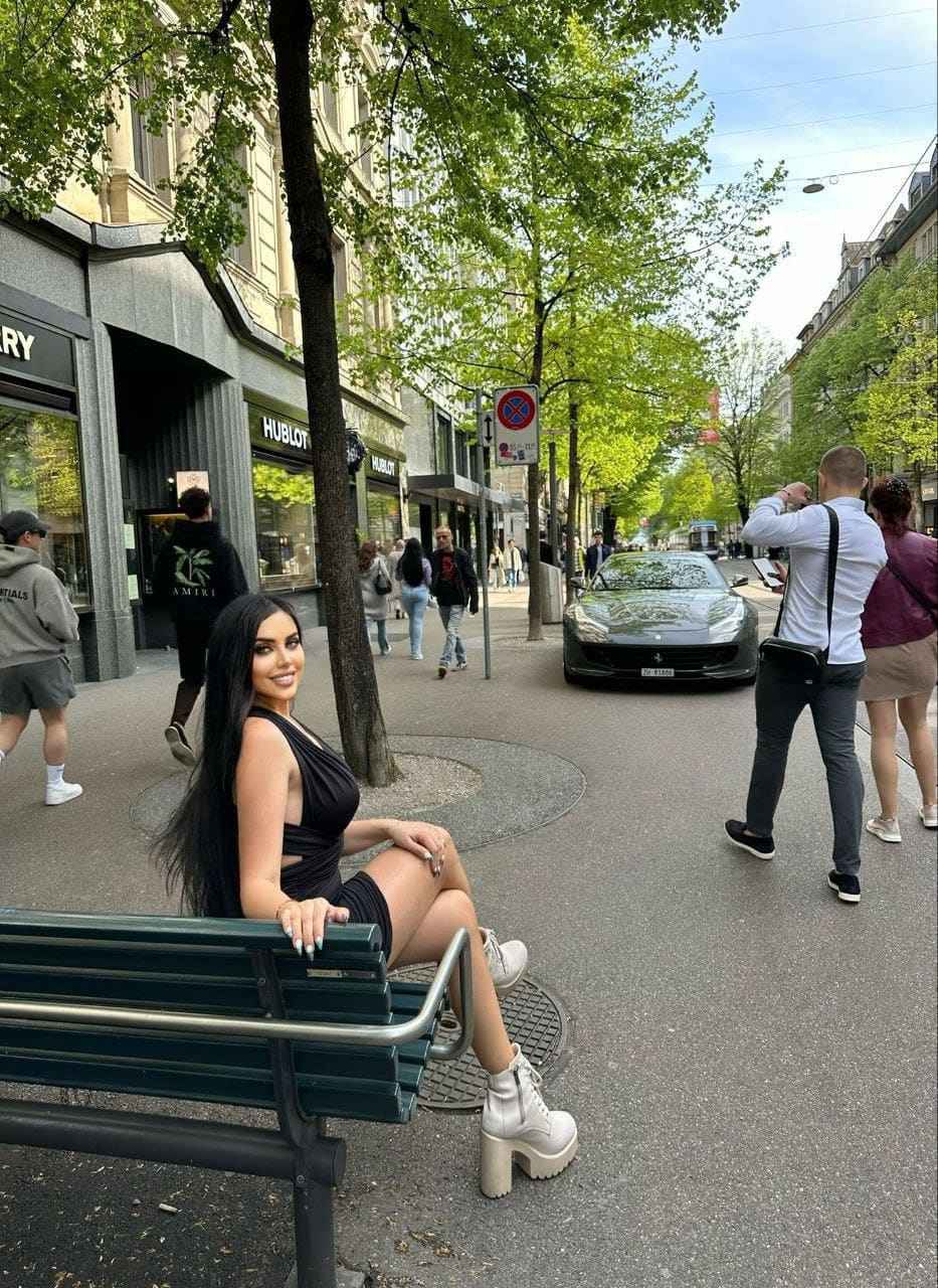 A woman in a black mini dress and white platform boots sits on a green city bench, with pedestrians and storefronts behind.