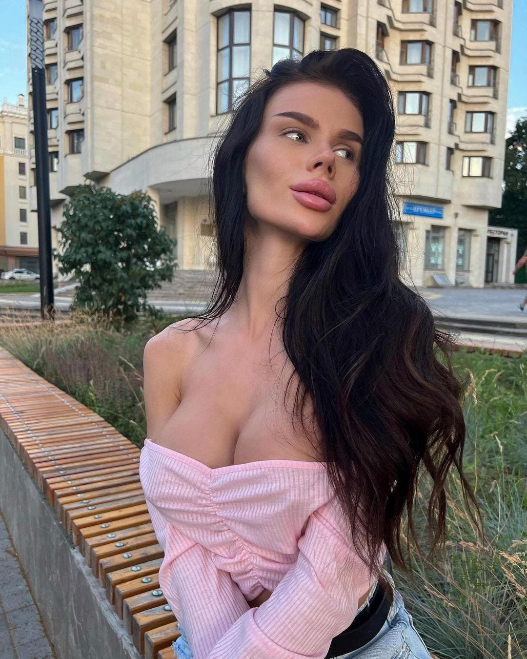 Outdoors before a large building, she wears a pink off-the-shoulder ribbed top and light pants, head tilted with eyes looking to the side.