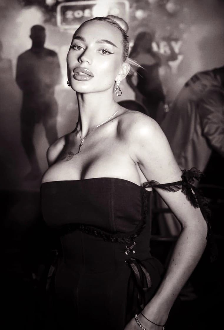 Woman in a black strapless lace-trimmed dress and jewelry, posing in a dimly lit indoor setting with blurred figures behind.