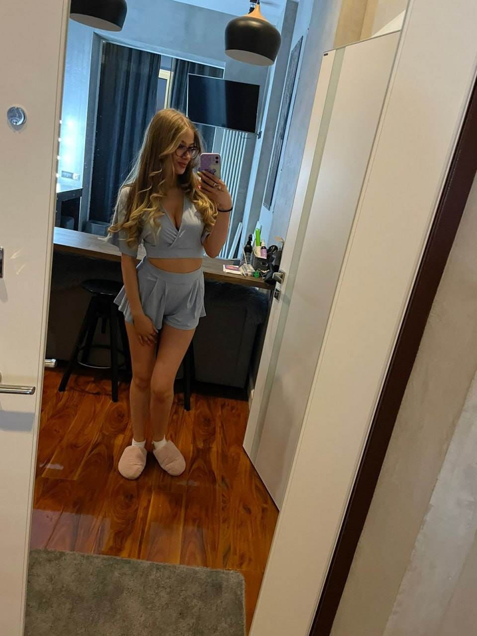 Mirror selfie in a grey crop top and shorts set, with glasses and white socks, in a room with wooden floors.