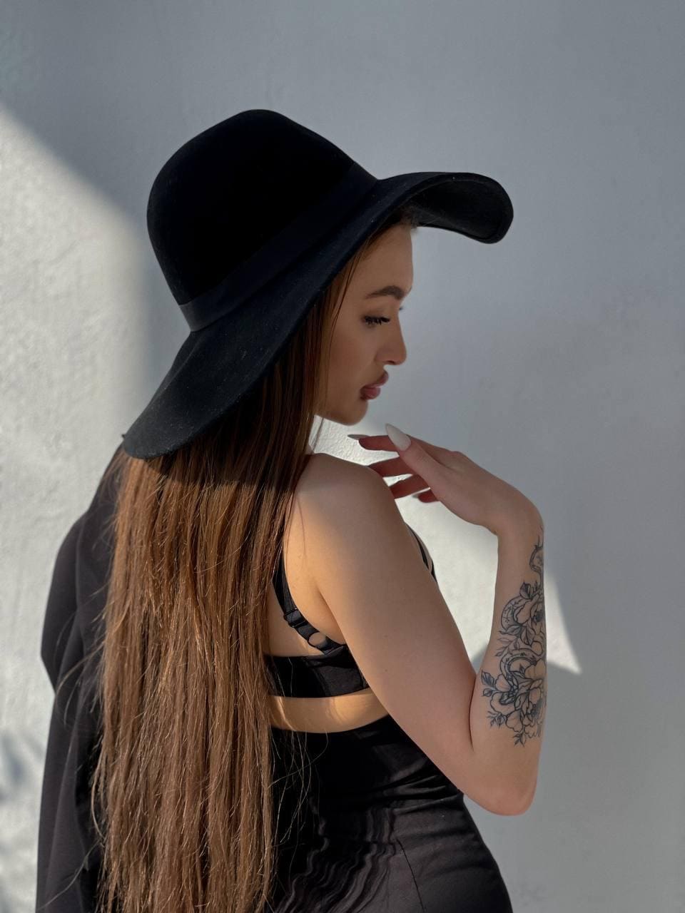 A woman in profile against a white wall wears a wide-brimmed black hat and open-back top, hand near her face, showing a floral forearm tattoo.