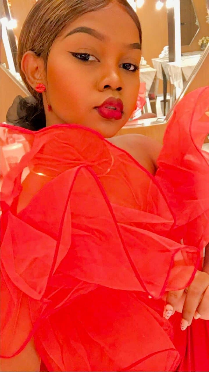 A woman with red makeup wears a sheer, ruffled red off-shoulder top in a warmly lit room with mirrors and tables.
