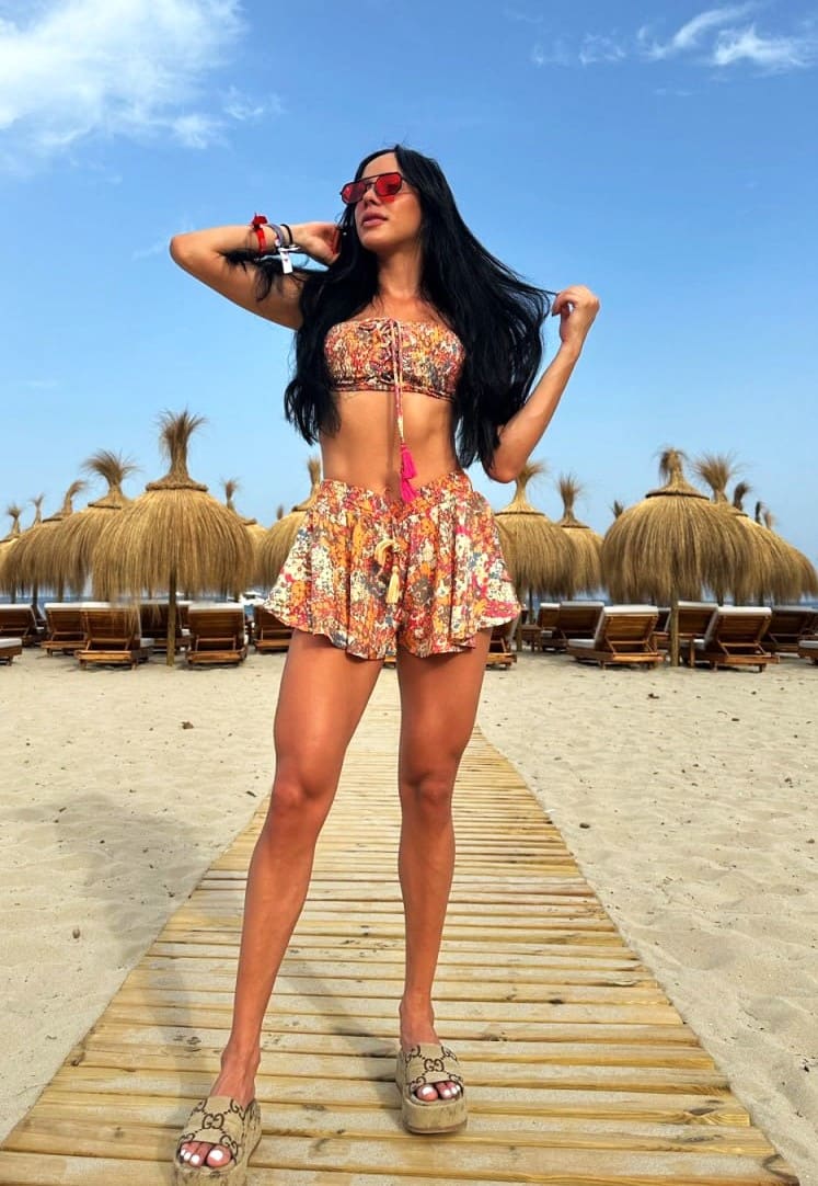 A woman stands on a beach boardwalk in a floral bandeau top and ruffled shorts, wearing red sunglasses and platform sandals near beach umbrellas.