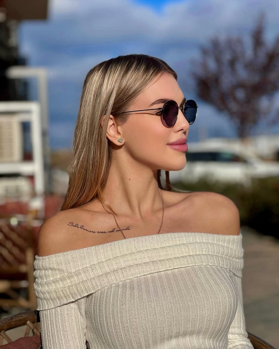 Seated outdoors with a blurred tree background, a woman in round sunglasses and a cream off-the-shoulder top looks toward the camera.