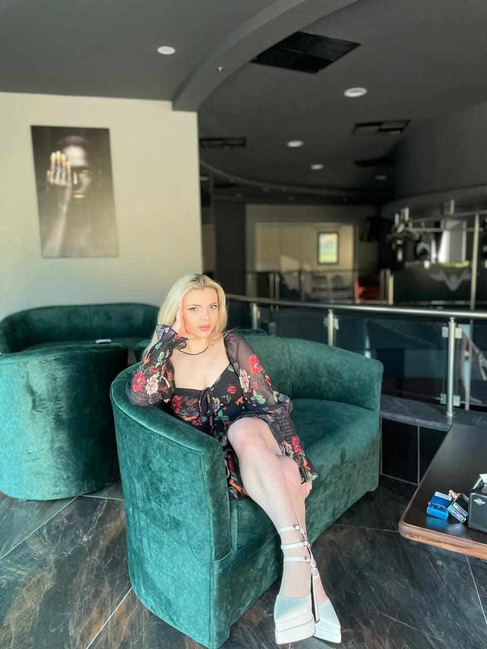 A blonde woman sits on a green velvet sofa in a sheer floral dress and white platform sandals, hand near her head.