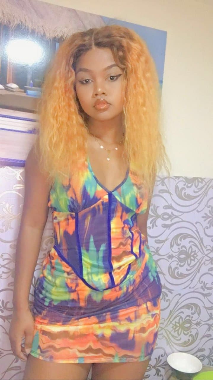 RUBY stands in a patterned mini dress with a deep V-neck against decorative wallpaper and a framed picture.
