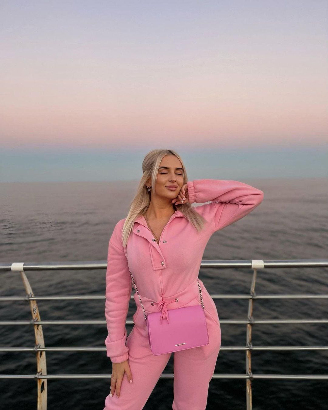 A blonde woman in a pink tracksuit stands on a ship deck, hand on hip, with open water and dusk sky behind her.
