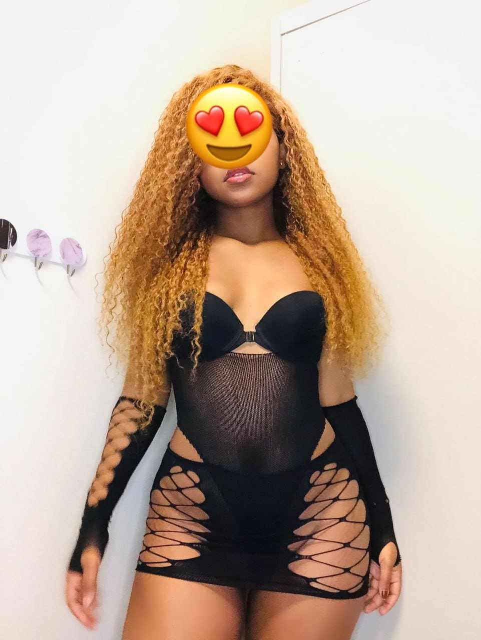 A blonde woman faces the camera in a black sheer mesh bodysuit with cut-outs and fingerless gloves against a white wall.