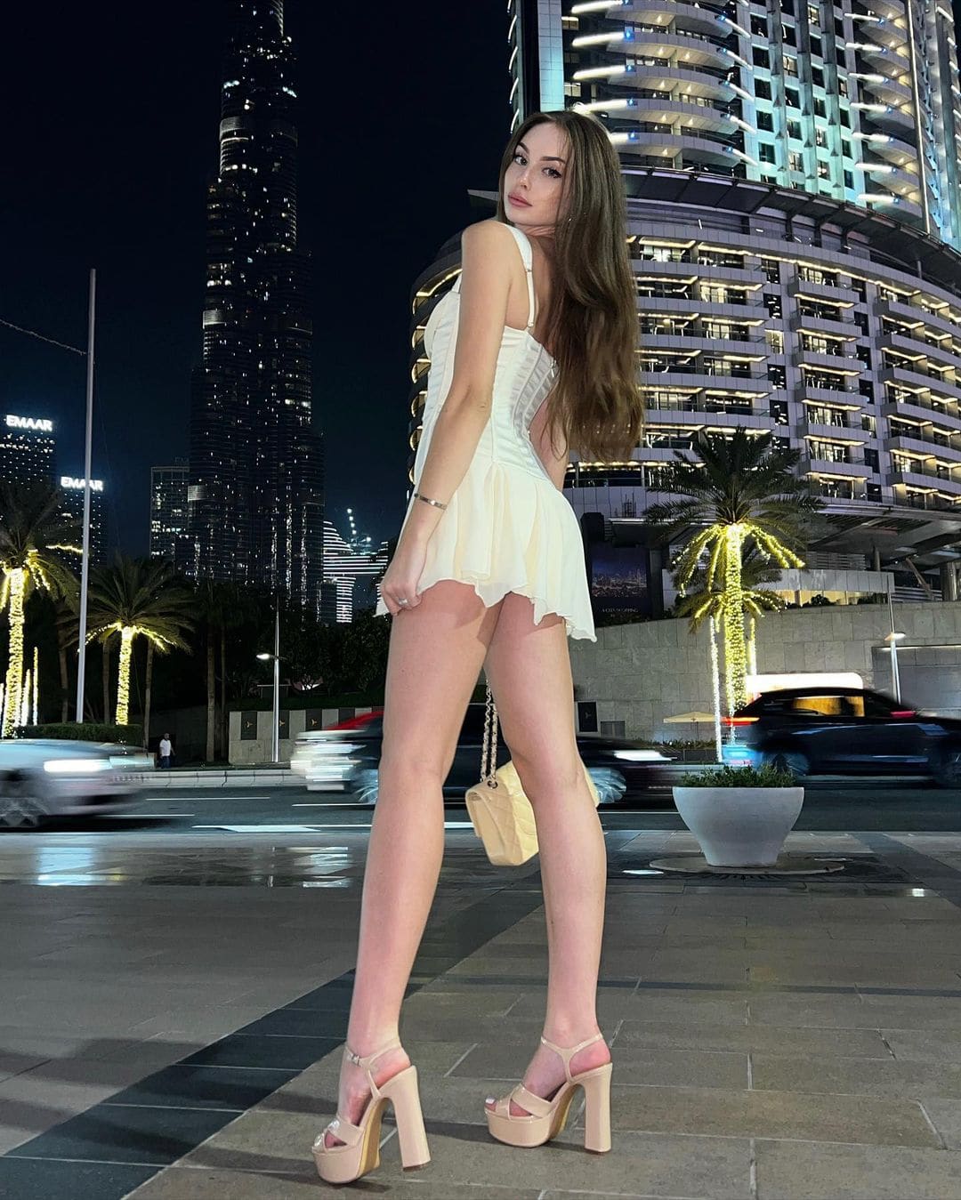 A woman in a white low-back dress and platform heels stands on a city sidewalk at night, with lit skyscrapers and palm trees.