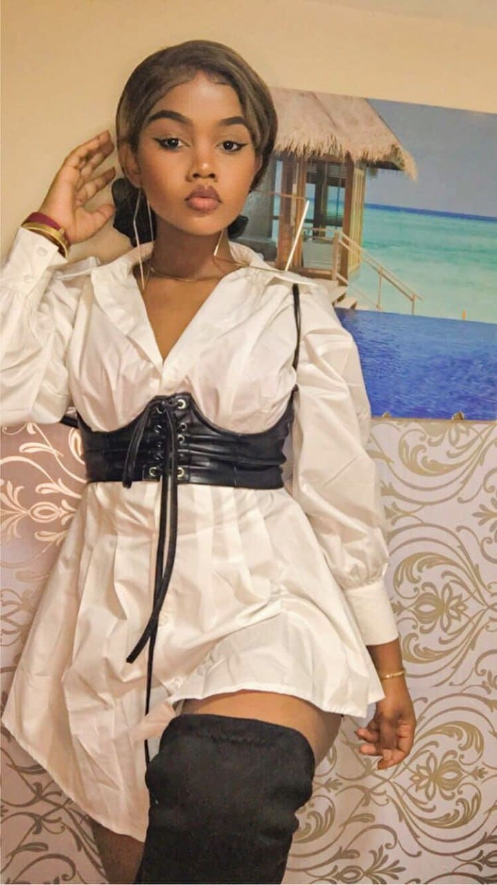 A woman in a white shirt dress, black corset belt, and thigh-high boots poses by a beach mural with one hand raised.