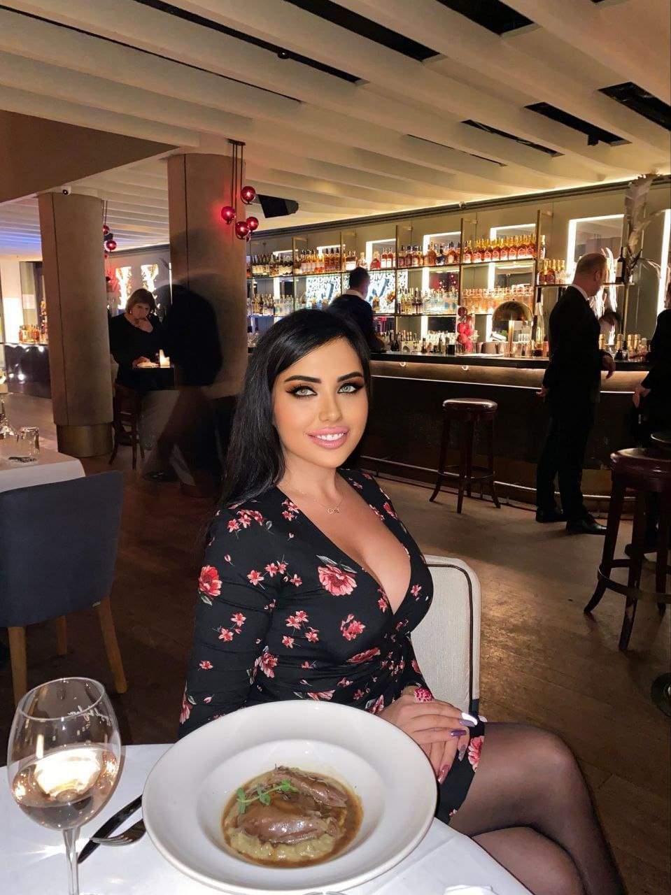A woman in a black floral dress and sheer tights sits at a restaurant table with food and wine, backed by a bottle-lined bar.
