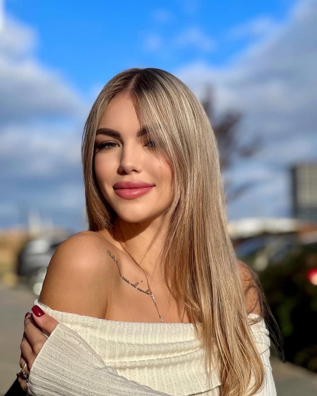 Sophia stands outdoors under a blue sky in a white off-the-shoulder top, one hand on her shoulder, revealing a collarbone tattoo.