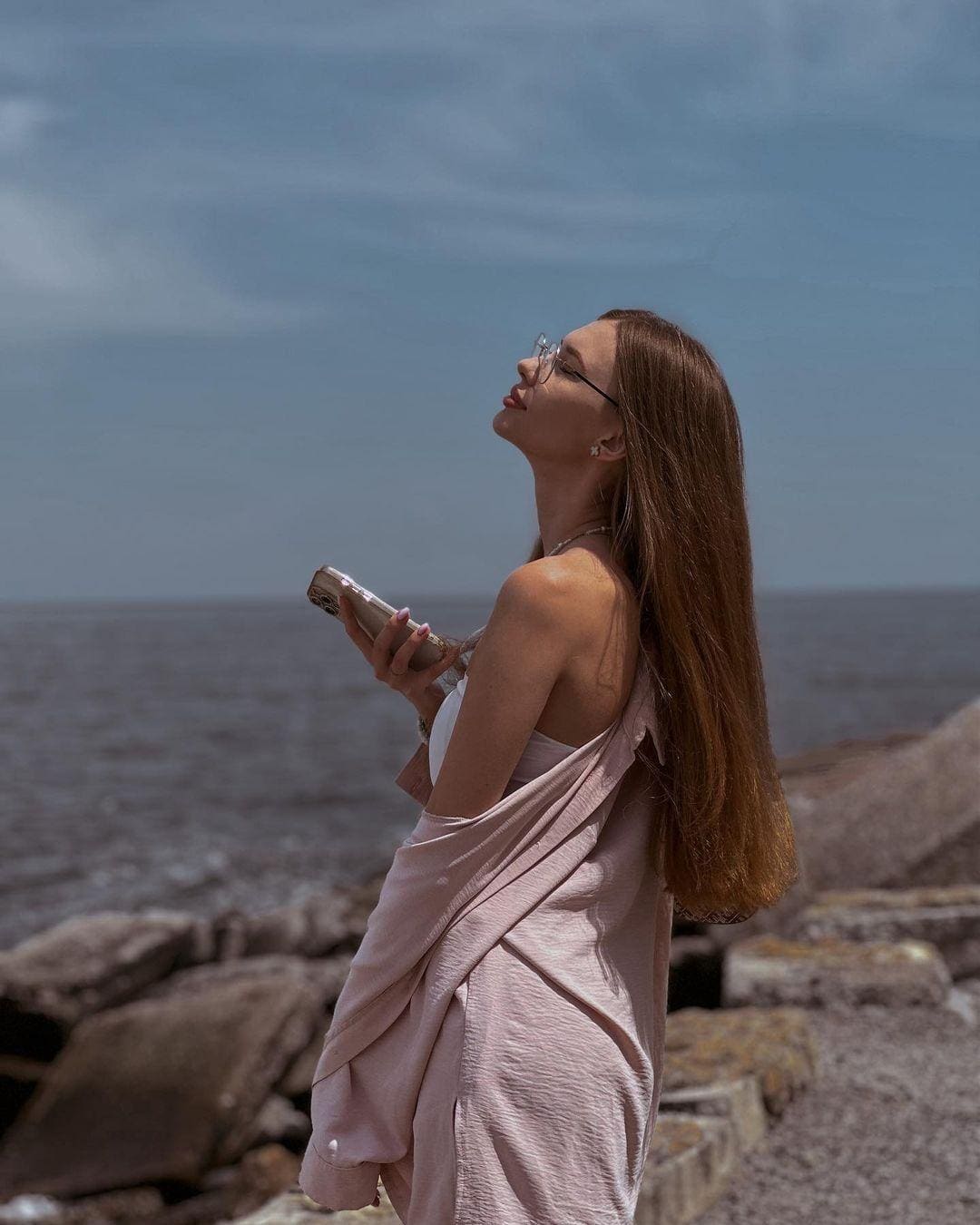 A woman in profile on a rocky shore wears a pink draped top and looks up while holding a phone against water and sky.