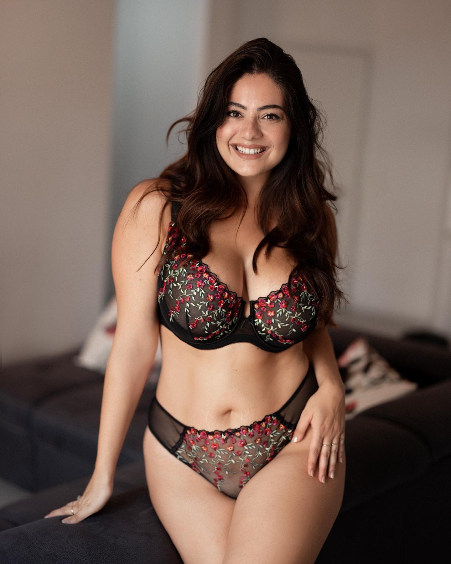 Celeste sits on a dark couch in a black and red floral lace bra and panties, smiling with one hand on her thigh.