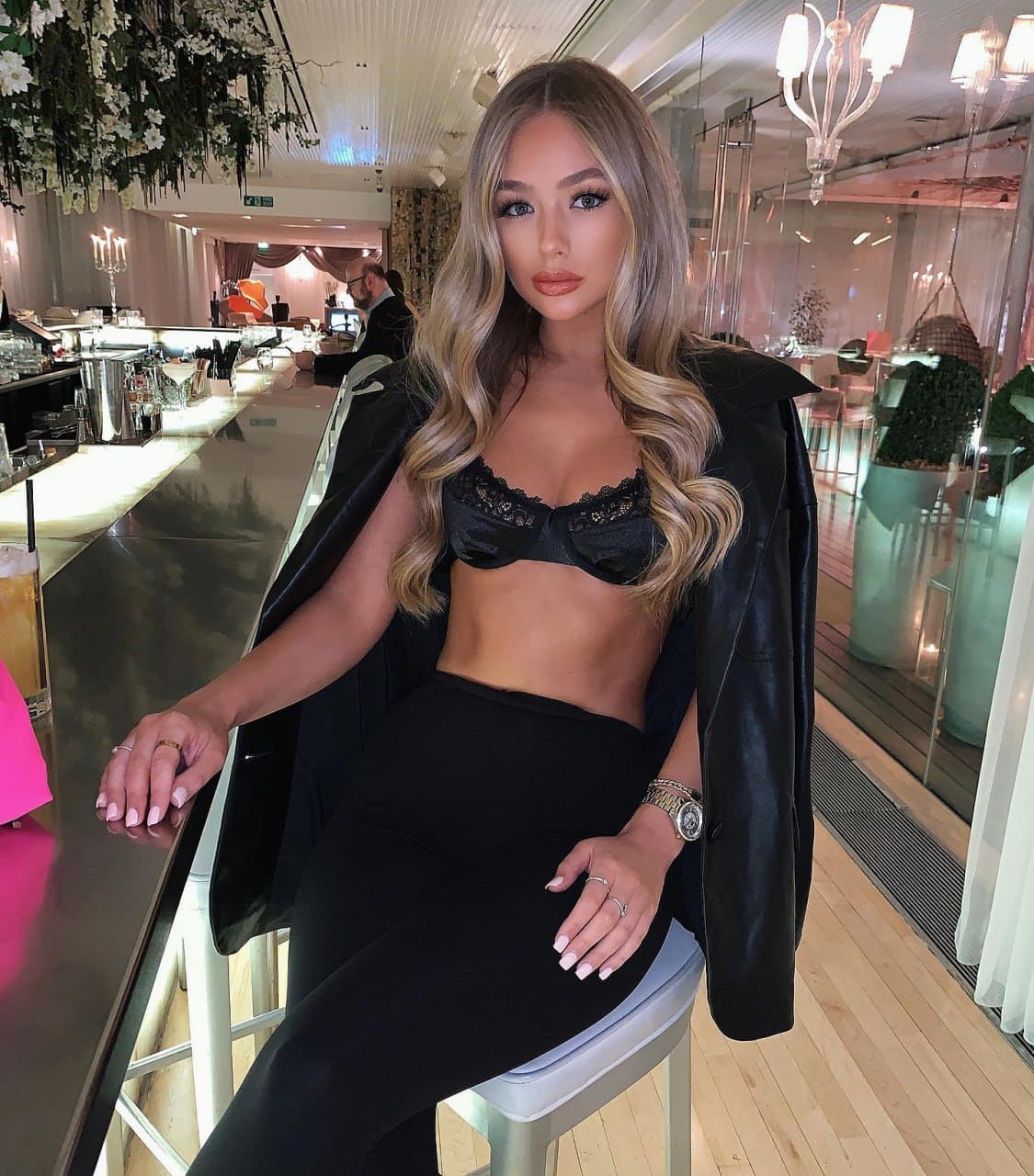Kalina sits on a white stool at a bar, wearing a black lace bra, high-waisted pants, and an open leather jacket in a lit venue.