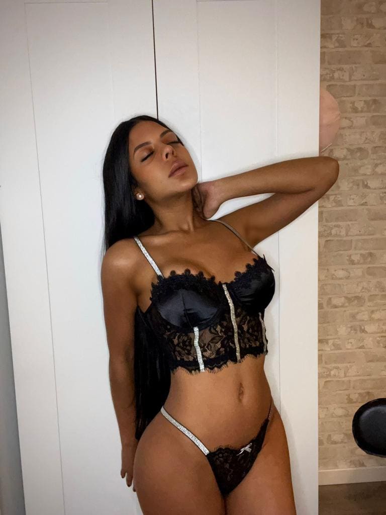 A woman in black lace lingerie with silver straps stands against a white and brick wall, arm behind her head, eyes closed.