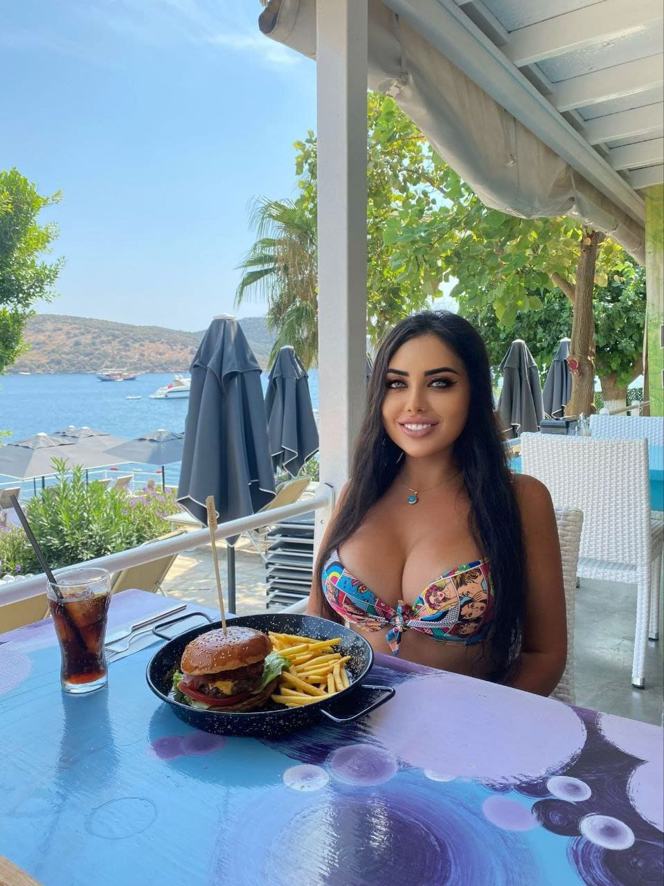 Nuri sits at an outdoor waterfront table with a burger and drink, wearing a colorful patterned bikini top near closed umbrellas.