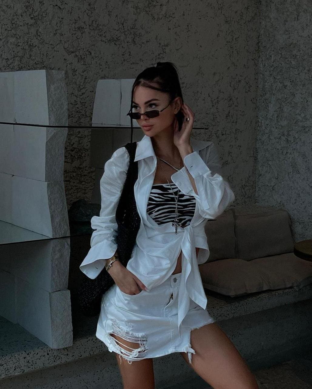 Woman with sunglasses, open white shirt over striped top, and distressed white mini skirt poses near a sofa.