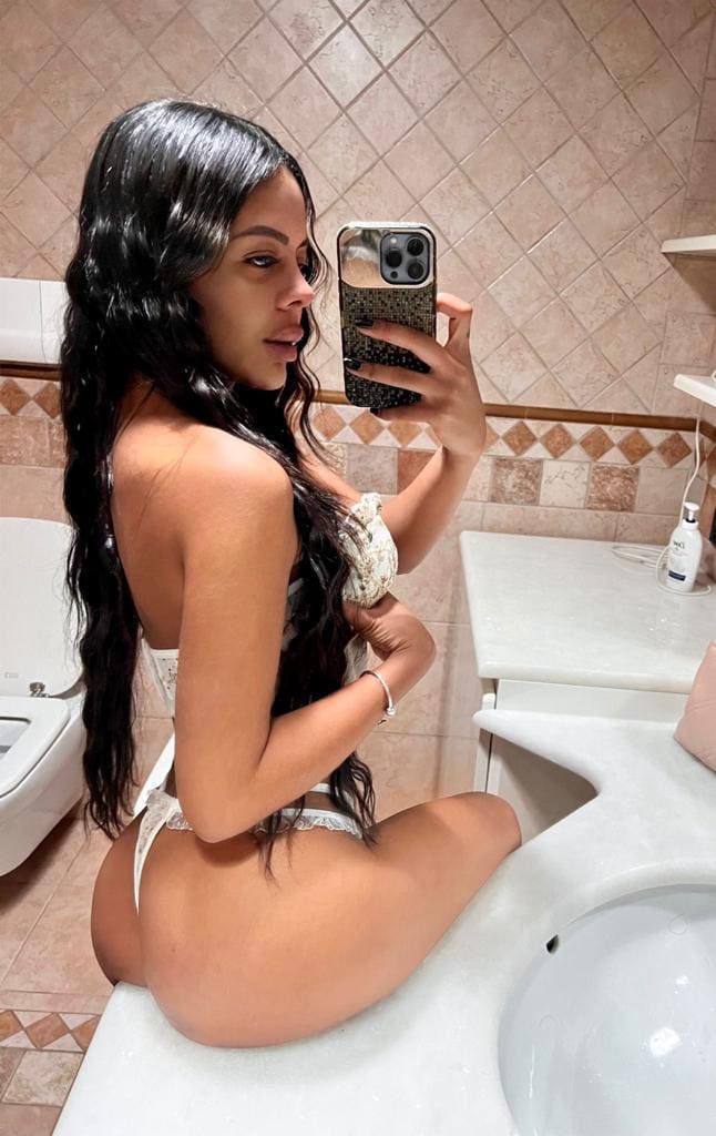 A woman in white lace lingerie sits on a bathroom counter taking a mirror selfie, with a tiled wall and sink in the background.