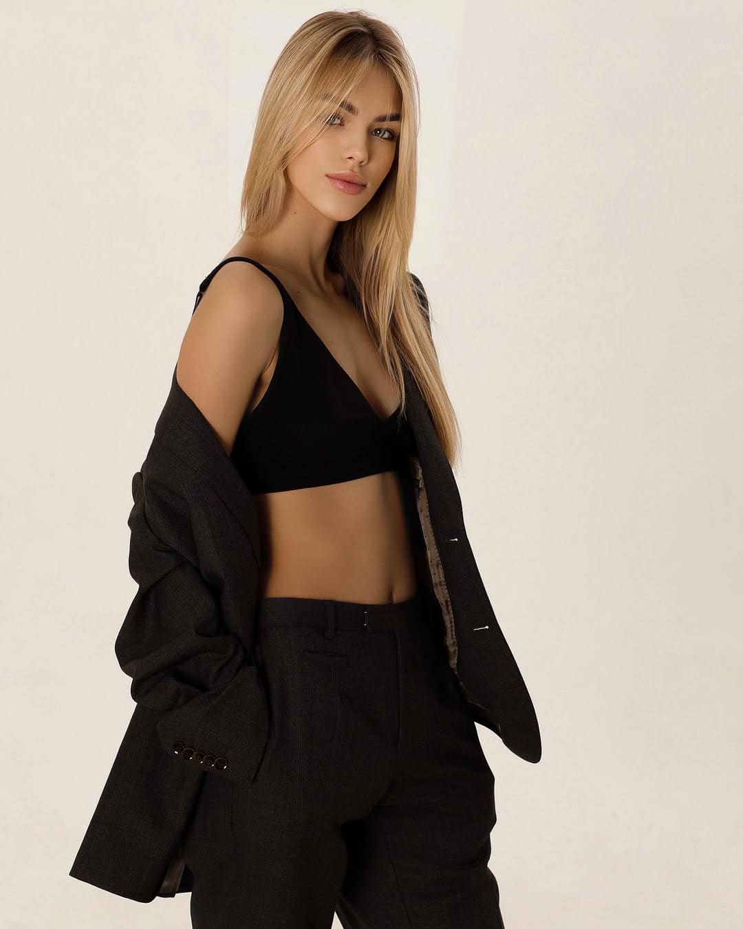 In a studio against a light background, a woman wears a black bralette, high-waisted trousers, and an off-shoulder blazer, hand in pocket.