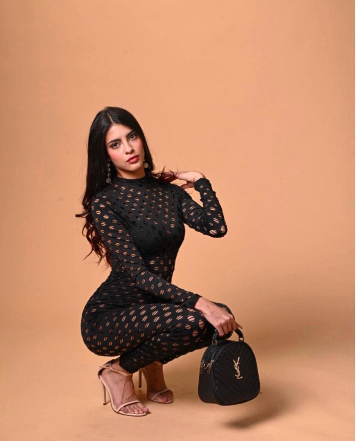 Crouching against a tan backdrop, she wears a black cut-out jumpsuit and nude heels, holding a black quilted handbag on the floor.