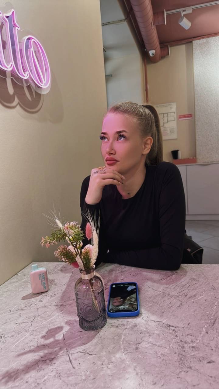 Seated at a marble table in a black long-sleeved top, resting chin on hand, with a vase and phone nearby.