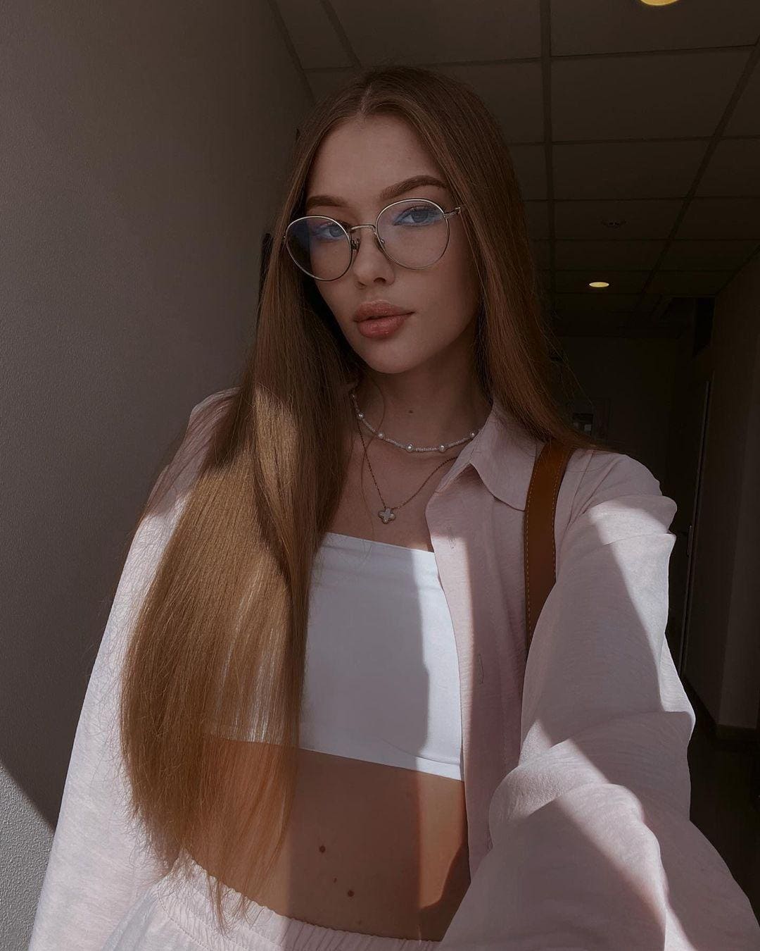 A woman with glasses and a pink shirt over a white bandeau top takes a hallway selfie, showing her midriff and necklaces.