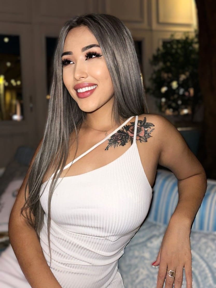 A woman with grey hair and a shoulder tattoo wears a white one-shoulder top in a room with a bed and potted plant.