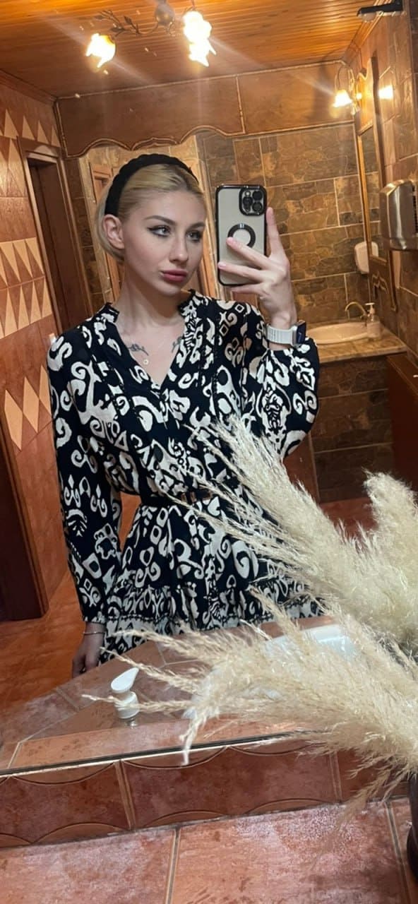 A blonde with a black headband takes a mirror selfie in a tiled bathroom, wearing a black-and-white patterned dress behind a counter with pampas grass.