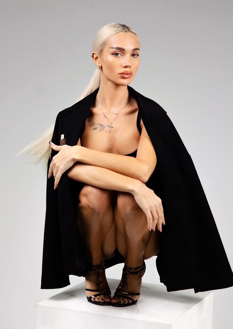 Blonde woman sitting on a white block in a black bra and coat, wearing strappy heels, with arms crossed over her knees.
