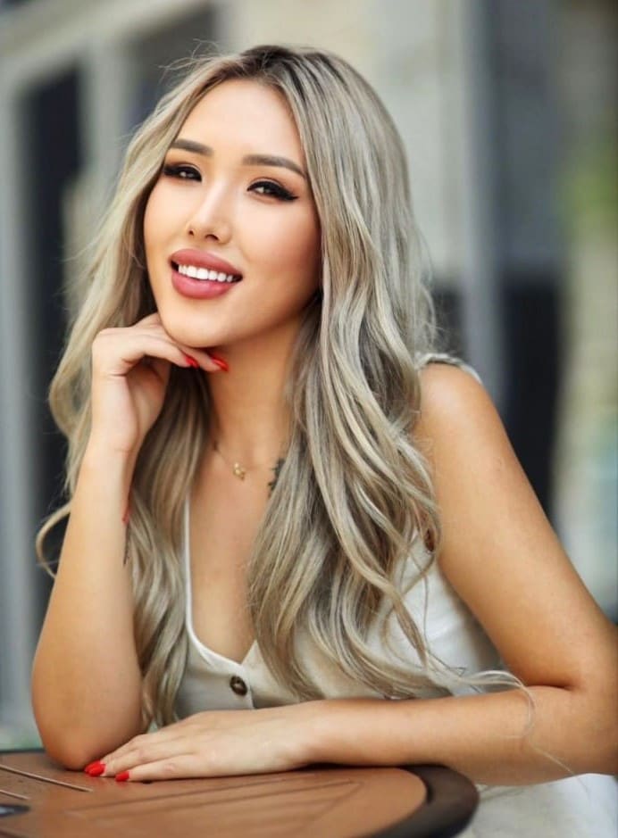 Anya rests her chin on her hand at a wooden table, wearing a white sleeveless top and necklace in a blurred outdoor setting.