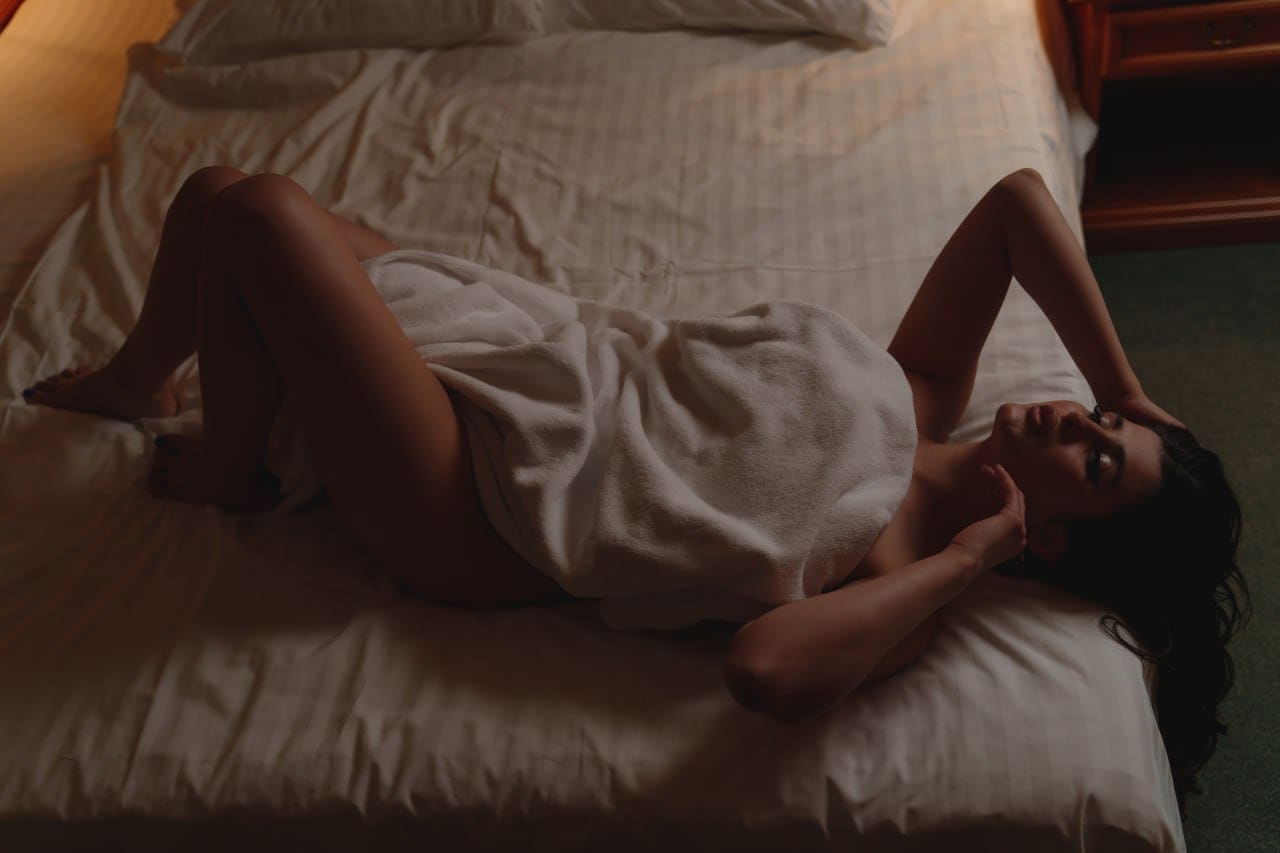 A woman lies on her side on a white bed, wrapped in a towel covering her torso, with one arm raised behind her head.