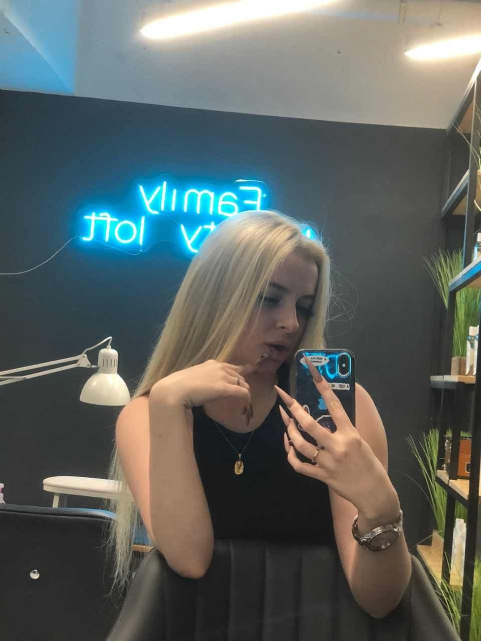 Woman taking a mirror selfie in a black top and dark skirt, touching her mouth near a blue neon sign.