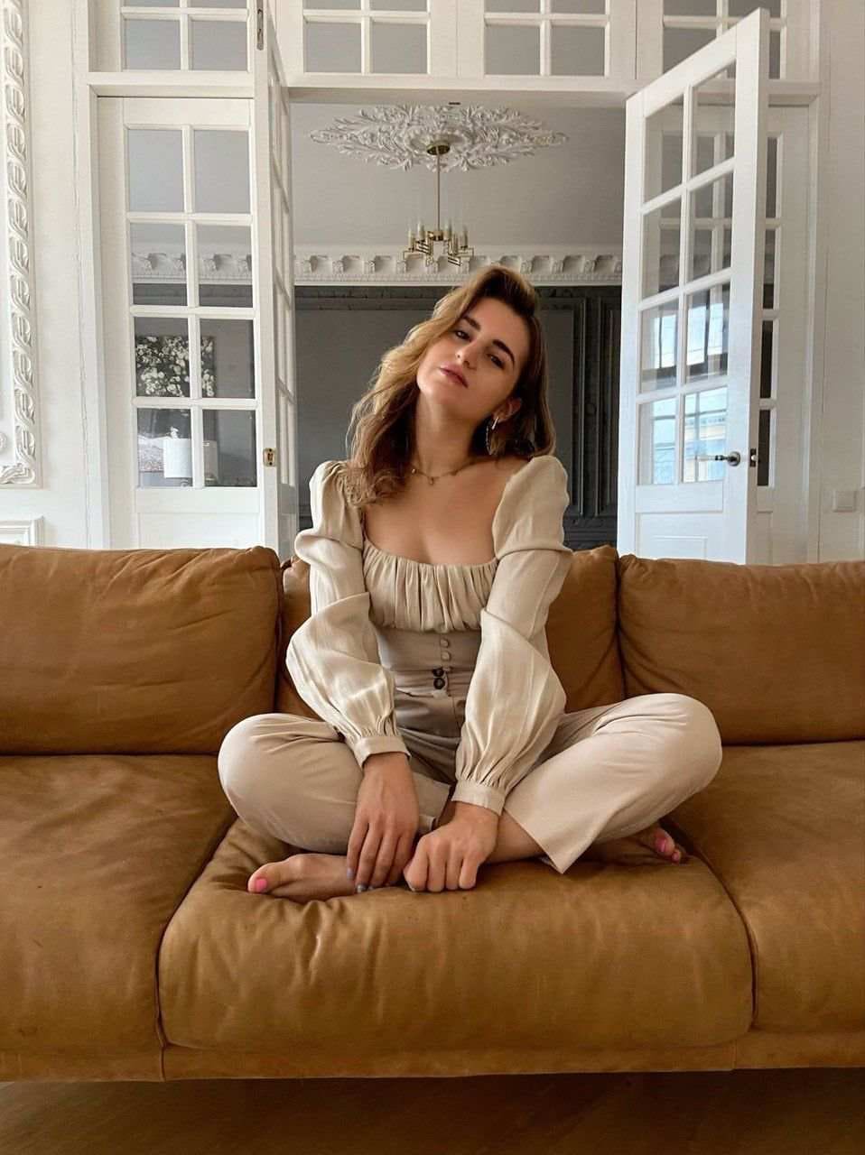 Christine sits cross-legged on a brown leather sofa in a cream top and pants, with white paneled doors and a chandelier behind her.