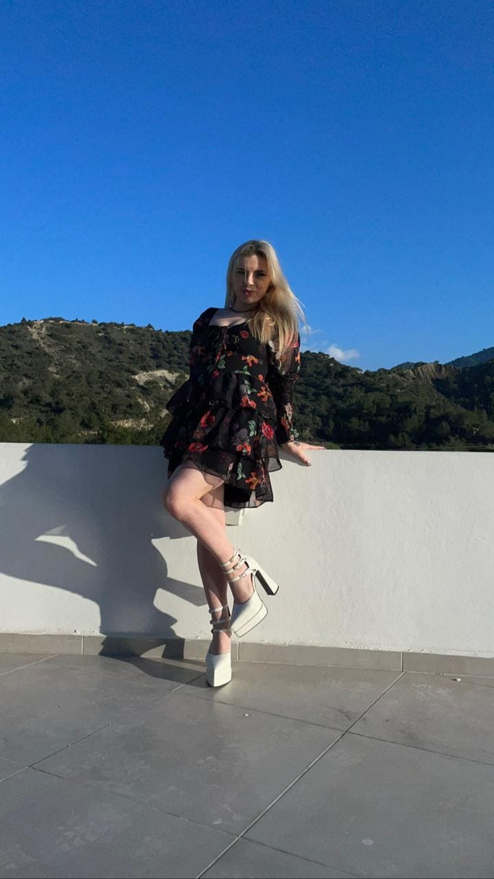 A blonde woman in a short black floral dress and white heels poses with one leg raised on a tiled terrace, hills in background.