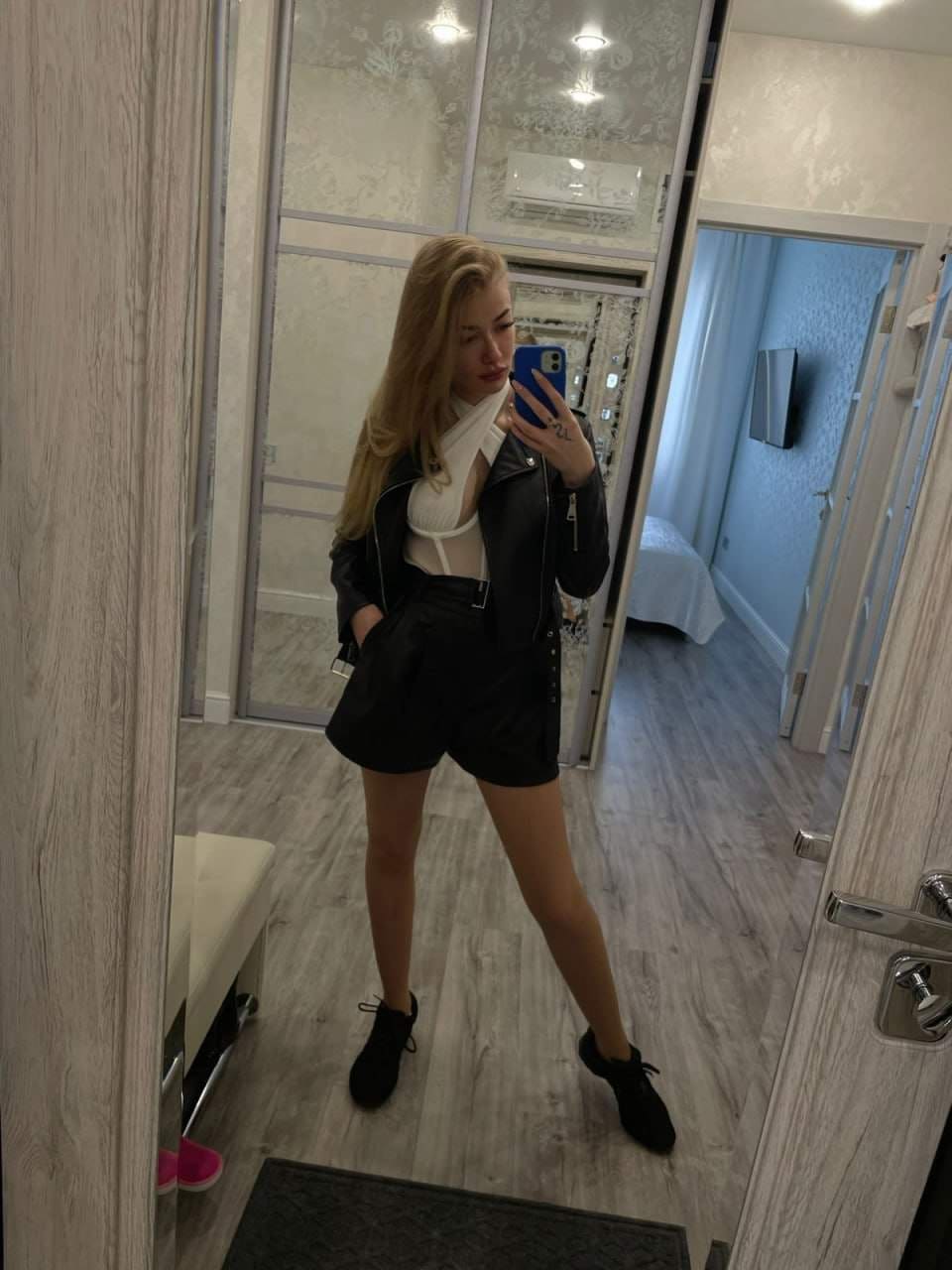 Mirror selfie in a bedroom showing a black leather jacket, white top, black shorts, and sneakers.