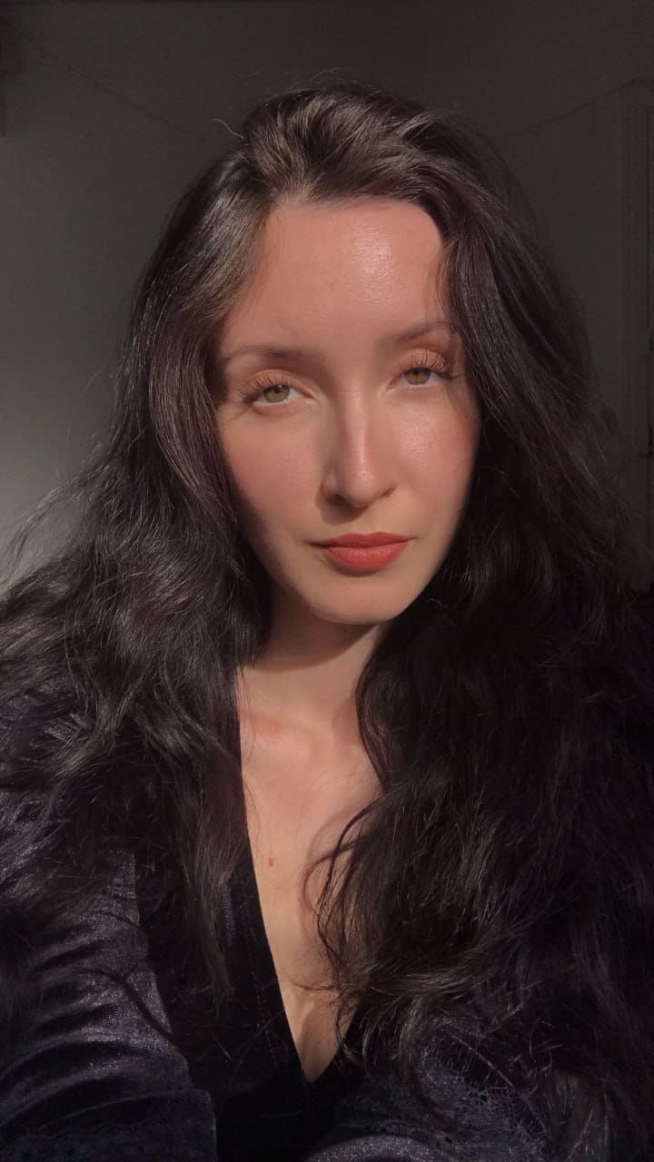 Thessa with long dark wavy hair wears a dark textured V-neck top against a dim, plain background.